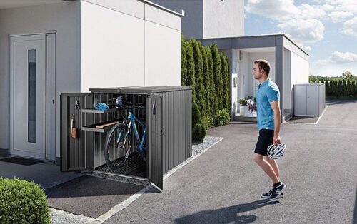 Abgebildet ist die Biohort MiniGarage. Sie ist geöffnet und steht neben einem Haus. Darin ist ein Fahrrad verstaut und ein Mann in einem blauen Tshirt steht daneben. Biohort Stauraum für den Garten.