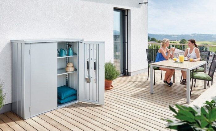 Silberner Terrassenschrank mit offen, stehender Tür. Terrassenschrank steht auf einem Holzbalkon vor einer weissen Wand.