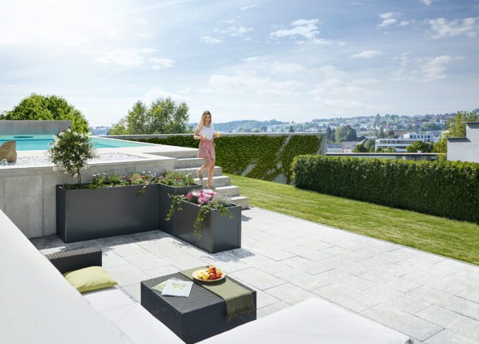 Das biohort DaVinci in dunkelgrau metallic auf einer grossen Terrasse mit Pool und Gartenlounge. Eine Frau steht auf der Treppe, die vom Pool zur Terrasse führt. Softub Schweiz