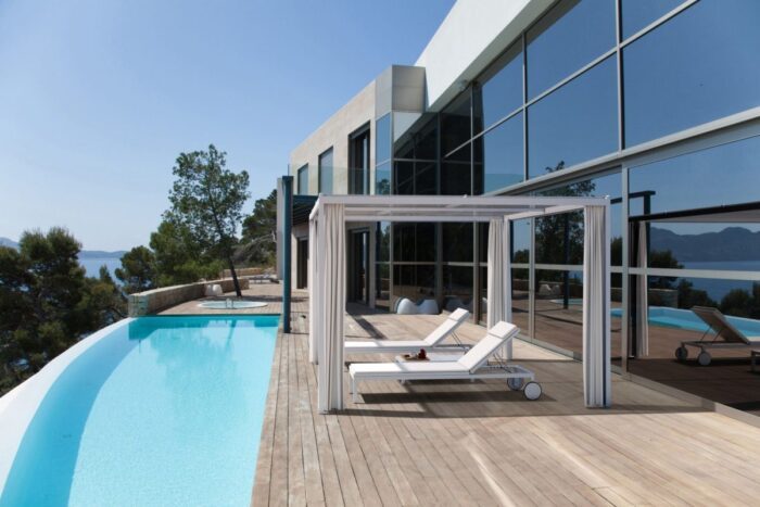 A beautiful pavilion with white bars and white deck chairs underneath. The pavilion stands infront of a big, modern house with a pool.